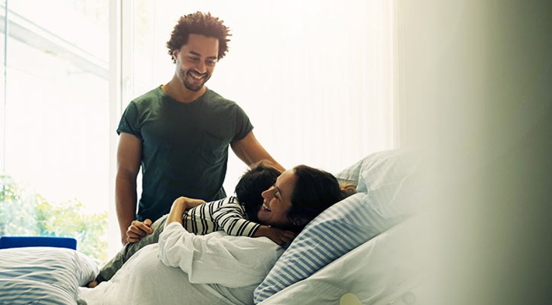 Frau liegt mit einem Kind im Bett und Mann steht daneben
