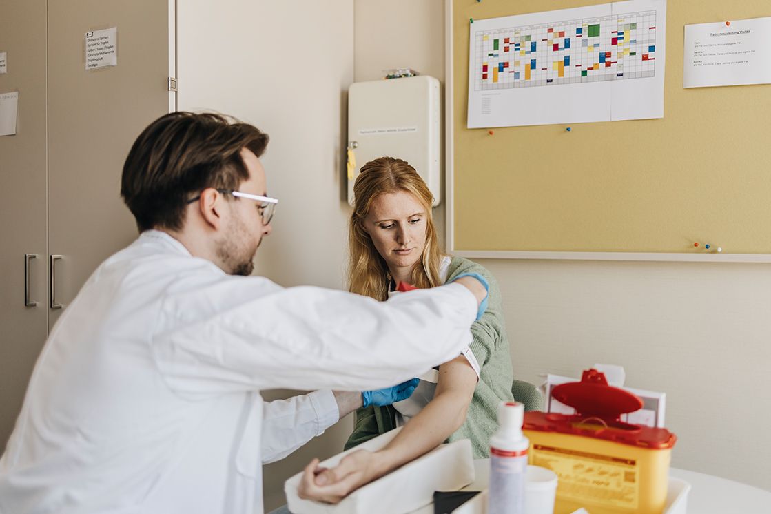 zwei Personen sitzen in einem medizinischen Behandlungsraum. Ein Arzt untersucht eine junge Frau und bereitet eine Blutentnahme vor