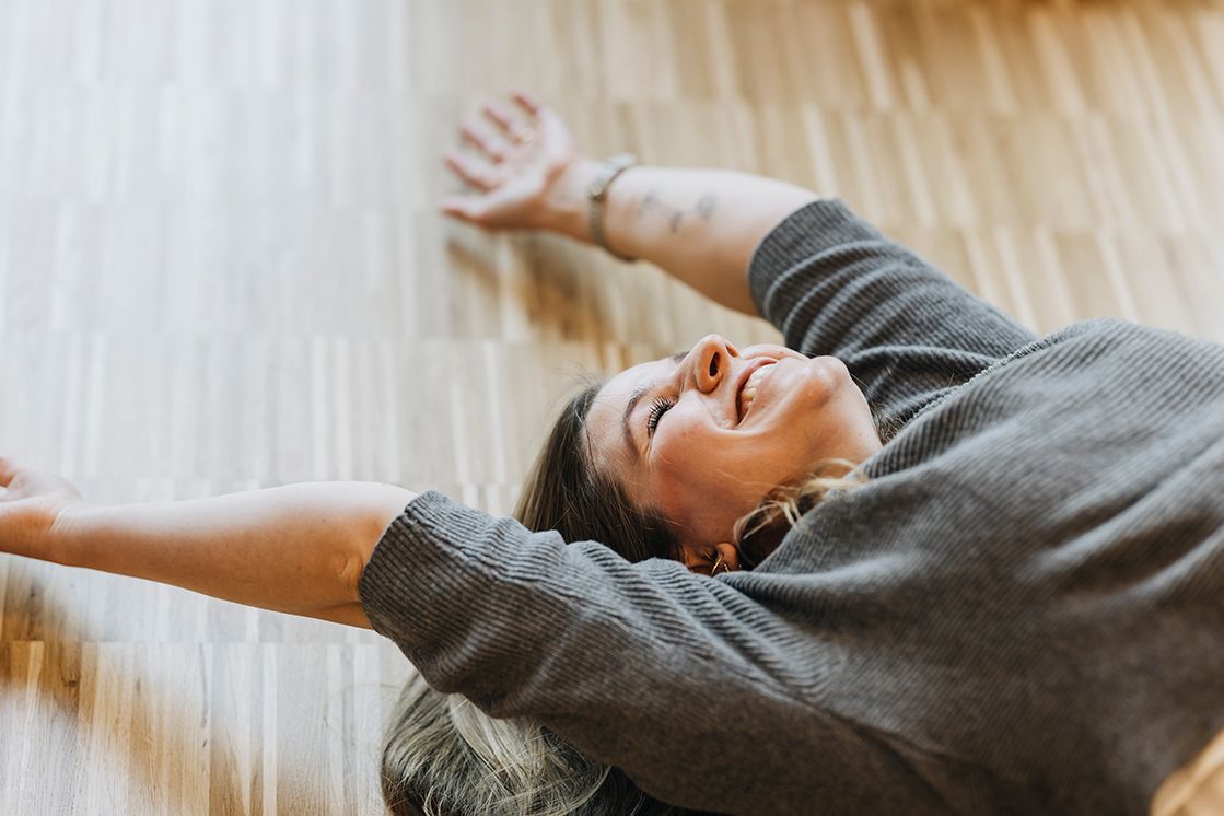 eine Frau liegt, beide Hände von sich gestreckt, stark Grinsend auf einem hellen Boden. Es wird lediglich der Oberkörper gezeigt.
