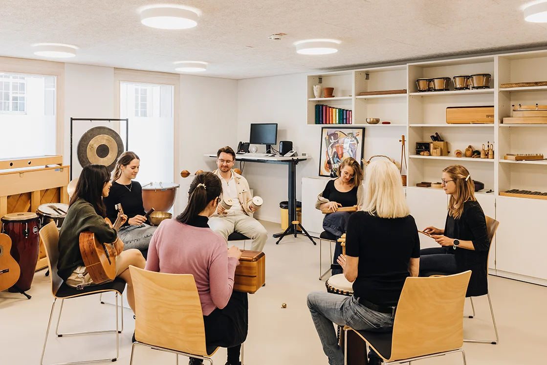 im Rahmen einer musiktherapeutsichen Sitzung, sitzen 7 Personen in einem Kreis und musizieren mit unterschiedlichen Instrumenten. In einem Raum voll mit anderen Instrumenten im Hintergrund