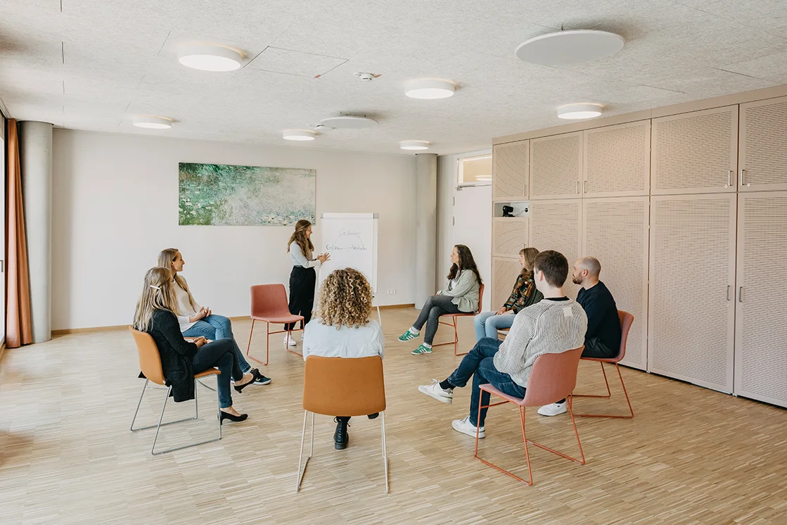 8 Personen sitzen auf Stühlen, in einem Sitzkreis und unterhalten sich in einem großen hellen Raum mit einem großen Bild an der Wand. Eine Person steht an einem Flipchart und erklärt etwas.