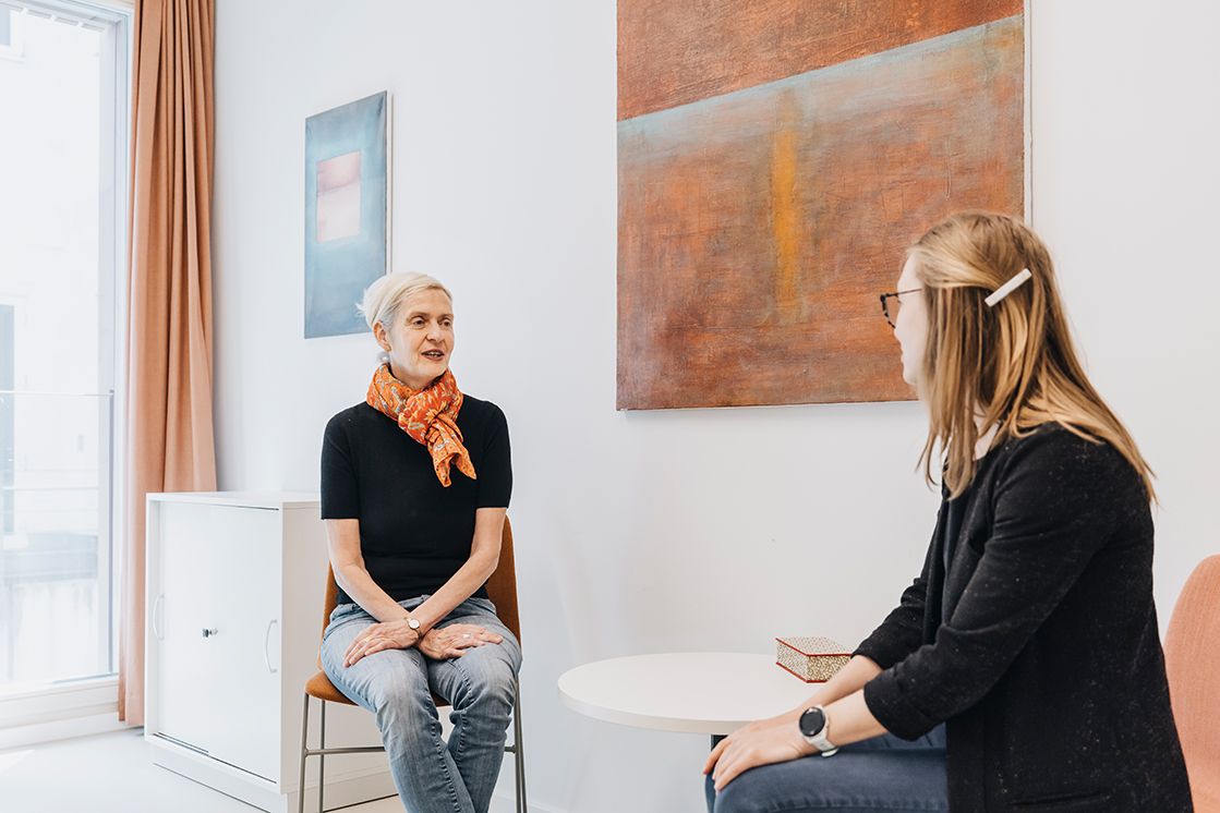 es sitzen sich zwei Frauen auf Stühlen, an einem Tisch gegenüber und führen ein freundliches Gespräch in Büroräumen mit bunten Bildern an der Wand