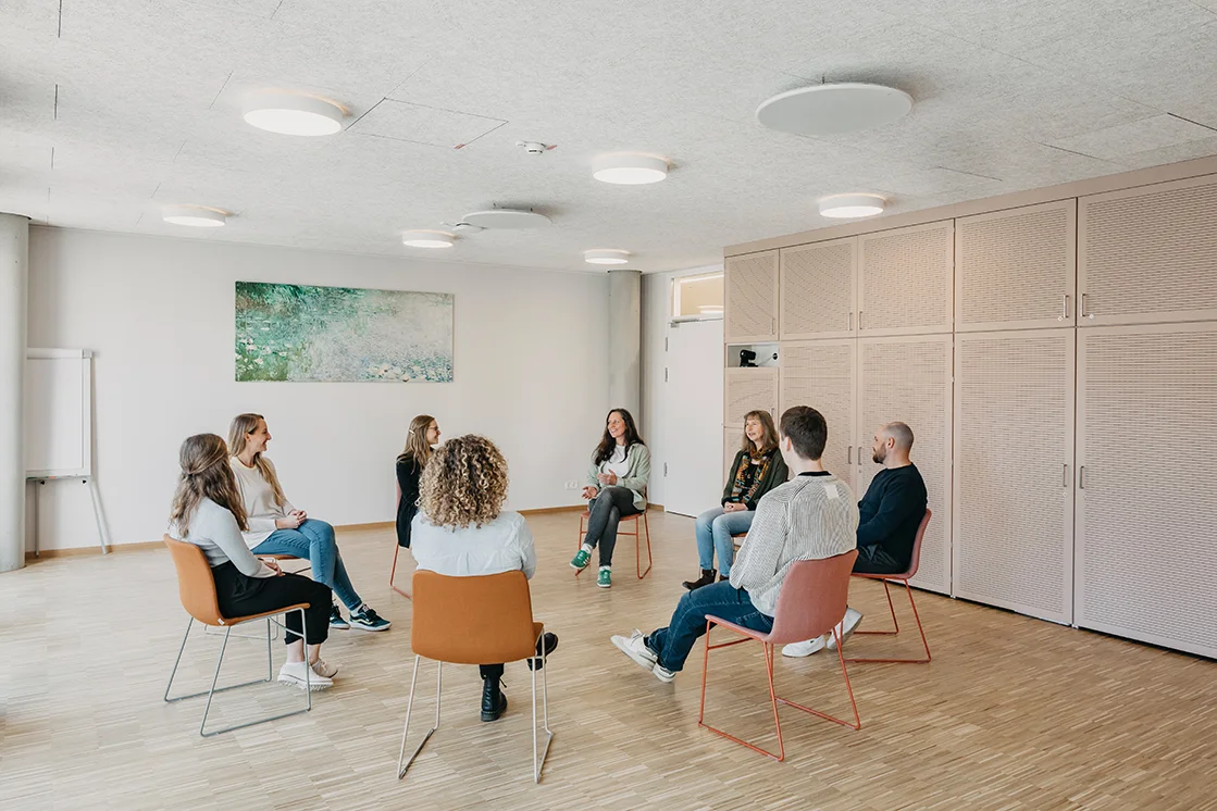 8 Personen sitzen auf Stühlen, in einem Sitzkreis und unterhalten sich in einem großen hellen Raum mit einem großen Bild an der Wand
