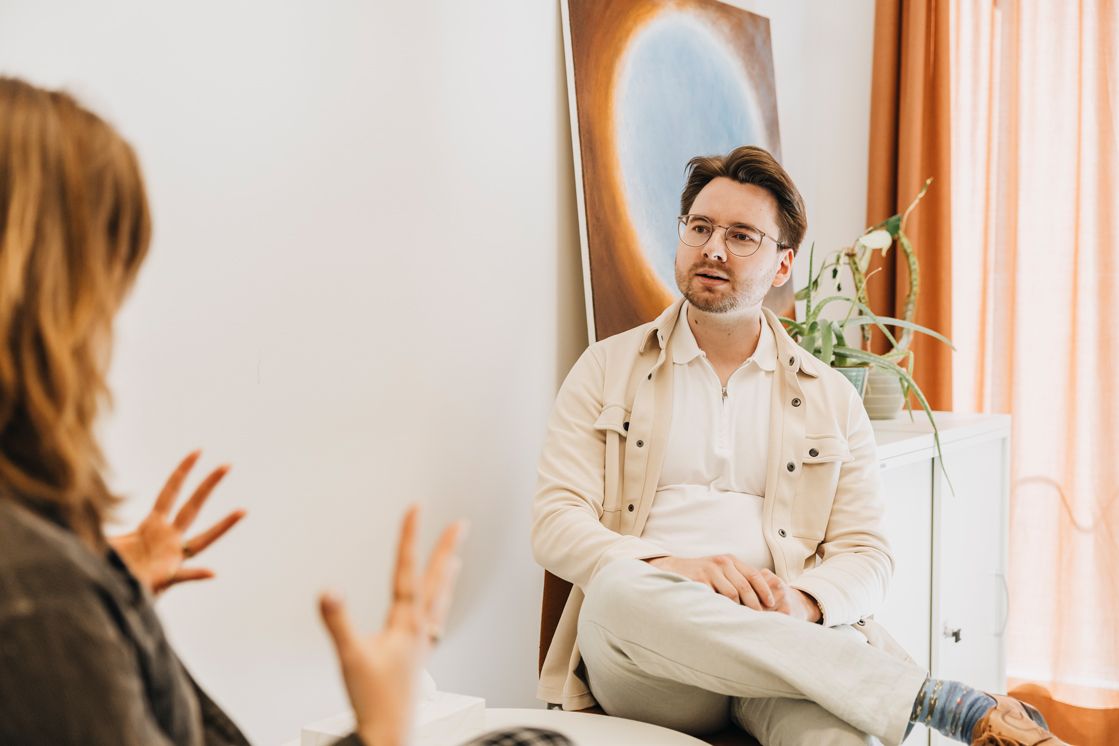 zwei Personen sitzen sich in einem Gespräch zugewandt gegenüber, in einem hellen Raum mit Sideboard und Pflanzen im Hintergrund