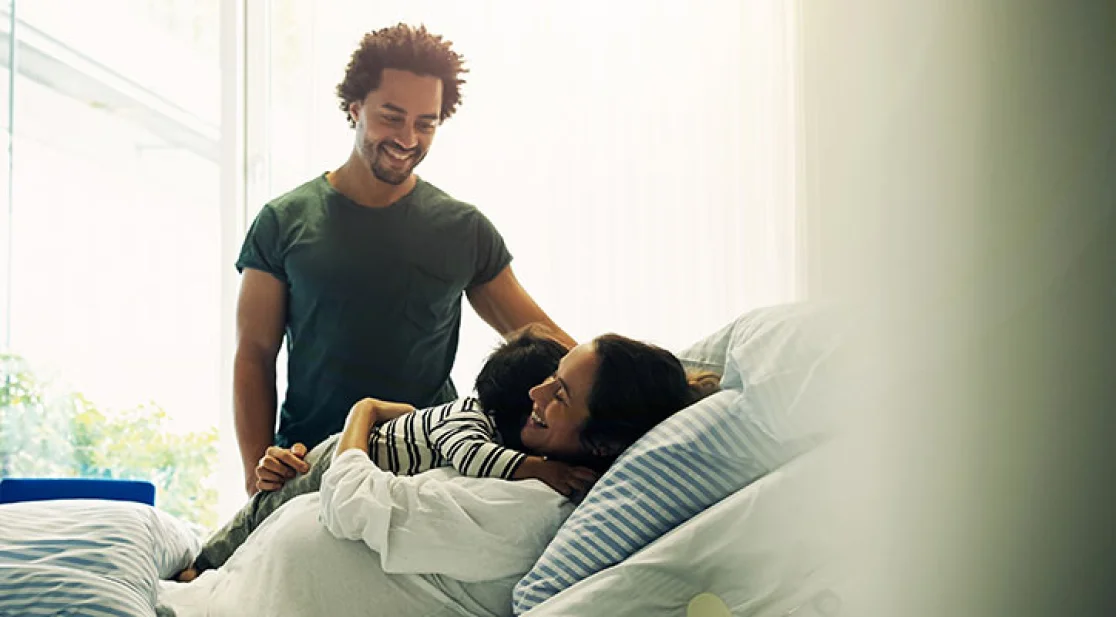 Frau liegt mit einem Kind im Bett und Mann steht daneben