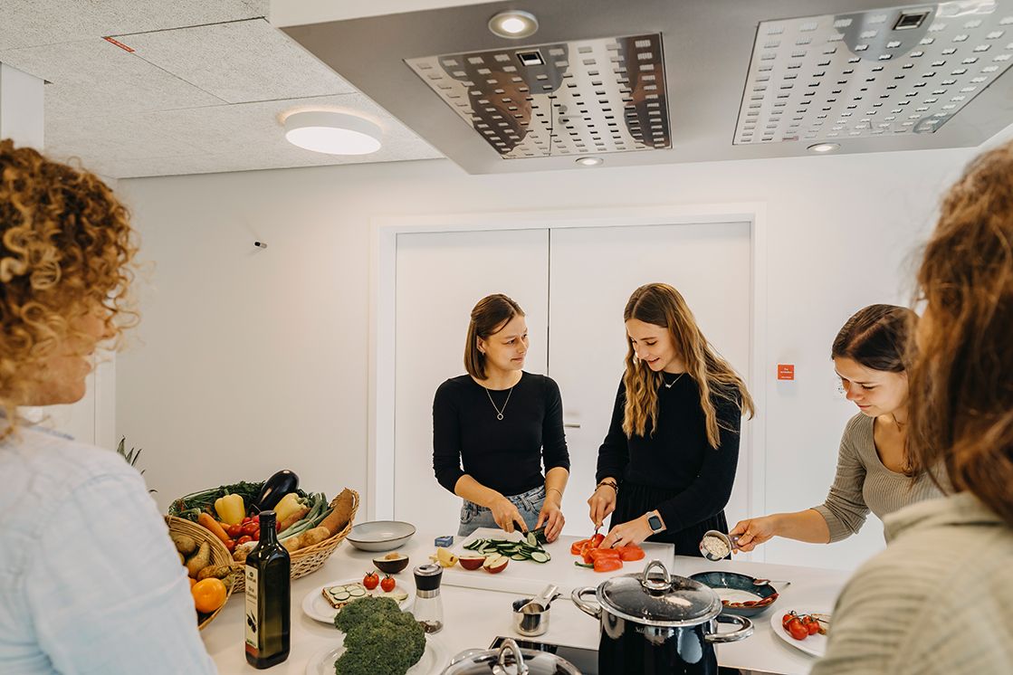 Es stehen 5 Frauen in der Küche und bereiten zusammen angeregt ein gesundes Gericht vor. In er Küche sind eine Vielzahl an Kochutensilien und Gemüse zu sehen.