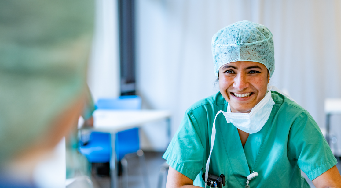 Eine Pflegekraft mit Haarnetz und zum Reden herabgezogenem Mundschutz im Gespräch mit einer Pflegekraft, die im Vordergrund  von hinten zu sehen ist