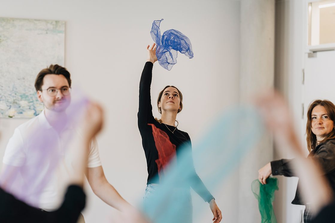mehre Personen werfen, in einem großen hellen Raum, Tücher in die Luft im Rahmen einer therapeutischen Sitzung. 