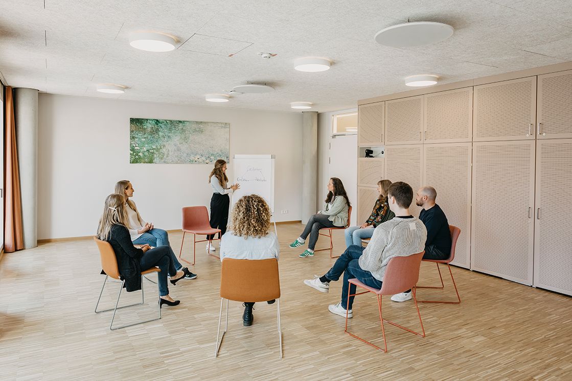 8 Personen sitzen auf Stühlen, in einem Sitzkreis und unterhalten sich in einem großen hellen Raum mit einem großen Bild an der Wand. Eine Person steht an einem Flipchart und erklärt etwas.