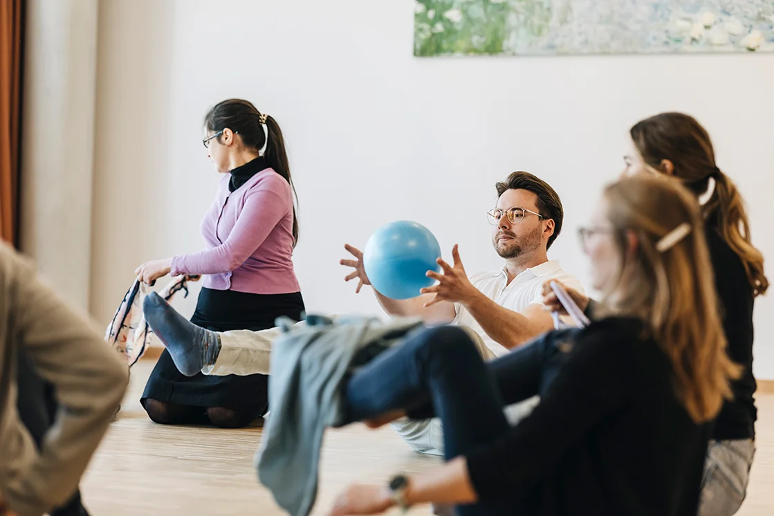 im Rahmen einer sportlichen Therapiesitzung, sitzen mehrere Personen auf dem Boden, eines hellen Raums. Einer der Personen führt eine Übung mit einem kleinen Ball durch