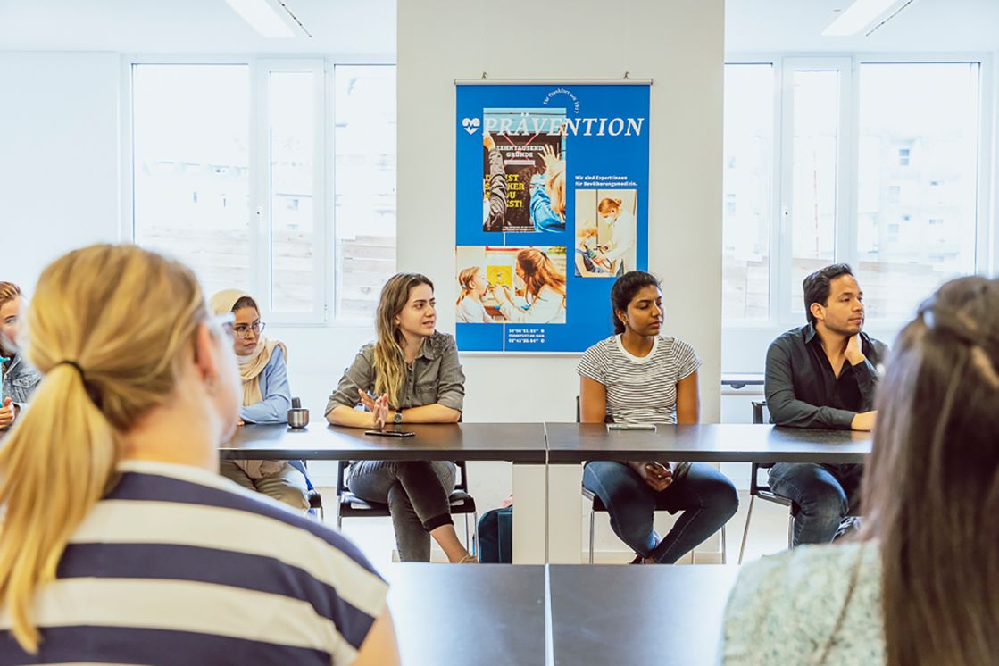 Studierende hören bei einem Vortrag zu und sitzen an Tischen