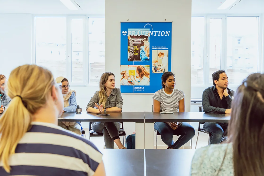 Studierende hören bei einem Vortrag zu und sitzen an Tischen