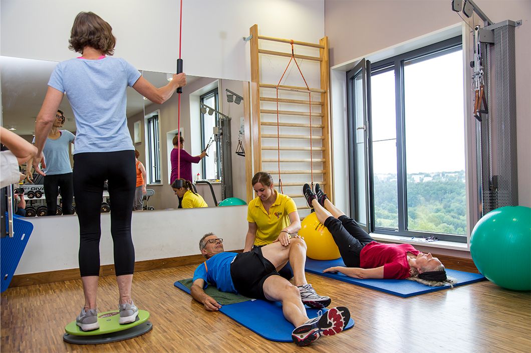 Personen trainieren unter Anleitung an verschiedenen Trainigsgeräten und auf Gymnastikmatten