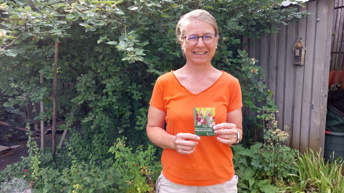 Ulrike Epp hält das neue Samentütchen in der Hand, im Hintergrund Garten mit Büschen, Stauden und einer Gartenlaube