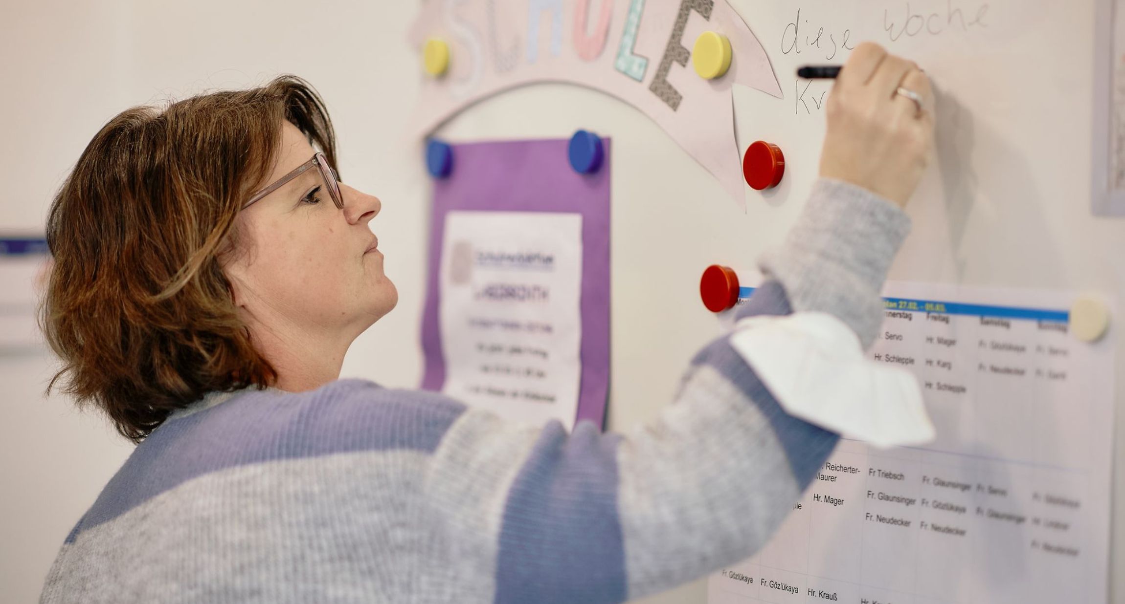 Eine Frau steht vor einer weißen Tafel. Sie trägt eine Brille und hat einen Arm erhoben, um mit einem Marker auf die Tafel zu schreiben. Vage ist im oberen Bereich das Wort "Schule" zu sehen. An ihrem Arm hängt eine FFP2-Maske.
