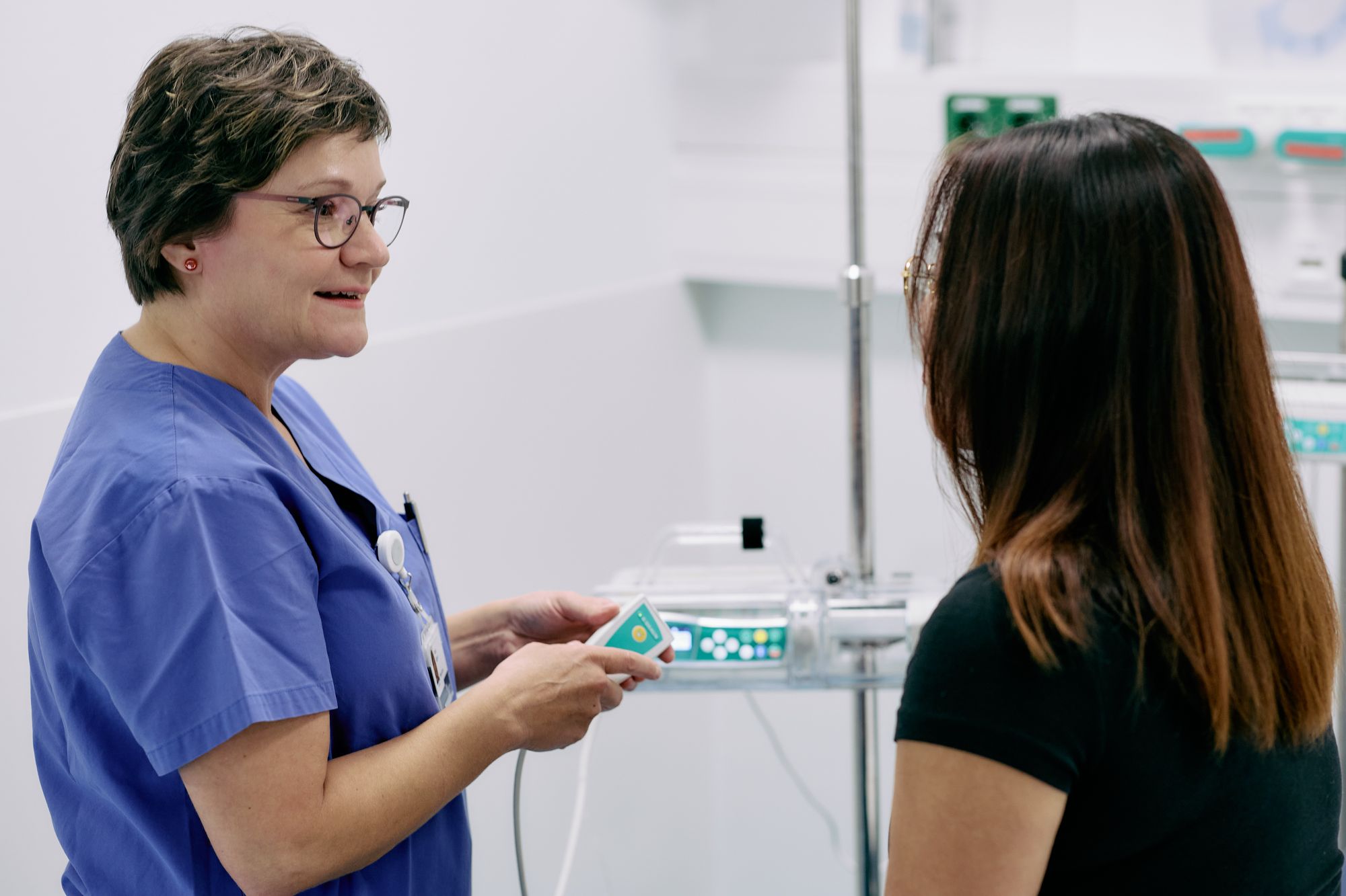 Ibolya Eggers, eine Pflegefachkraft in blauem Kasack, mit Brille und kurzen dunklen Haaren steht im Gespräch mit einer Patientin. Die Patientin ist nur von hinten zu sehen.