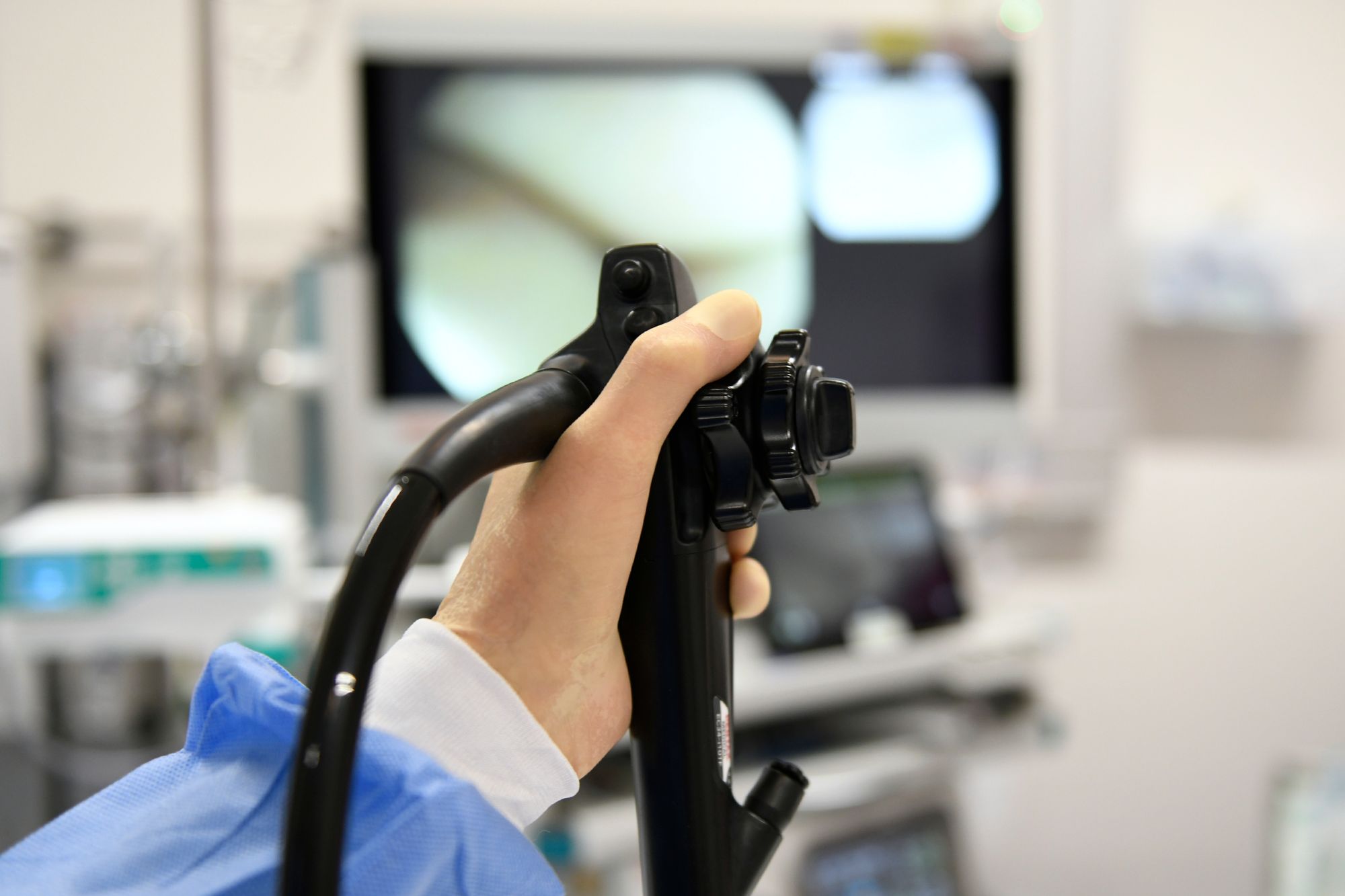 Eine Hand hält das Bedienelement eines Edoskops. Im Hintergrund ist verschwommen ein Krankenhauszimmer mit Bildschirmen zu erkennen.