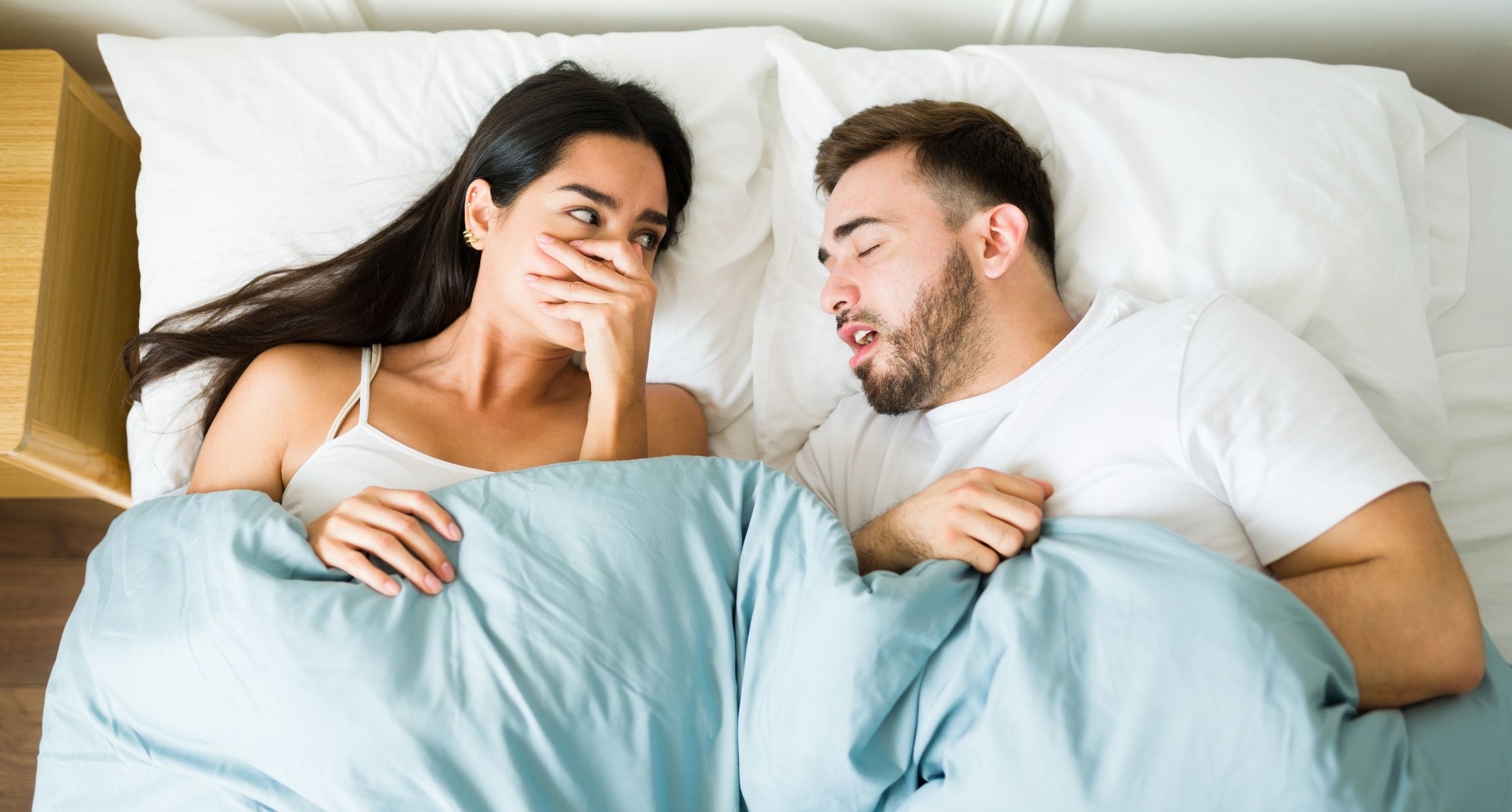 Ein Paar liegt morgens im Bett, er atmet noch schlafend in ihre Richtung und sie hält sich die Näse zu.