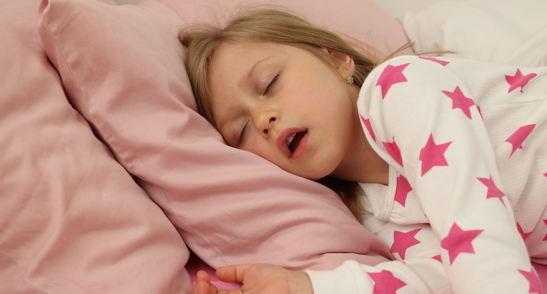Ein Mädchen in einem weißen Schlafanzuzg mit pinken Sternen liegt mit offenem Mund im Bett.