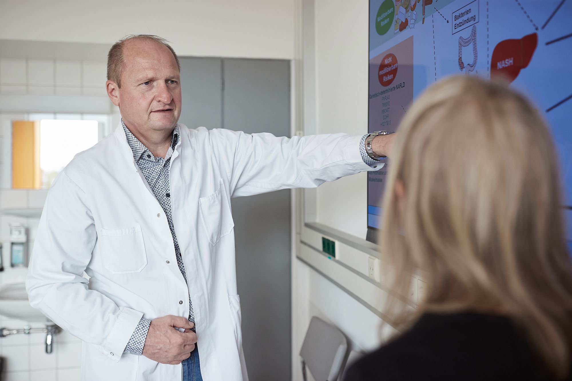 Prof. Norbert Stefan, ein Mann im Arztkittel, steht vor einem Bildschirm und erklärt einer Patientin eine Darstellung. Die Patientin ist nur von hinten verschwommen zu erkennen.